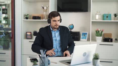 Portrait-of-a-young,-handsome,-and-cheerful-man,-listening-to-music-on-wireless-headphones-while-sitting-at-a-desk-in-a-bright-office-wearing-business-attire