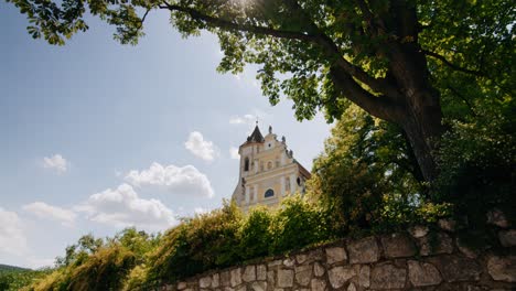 Catholic-Church-Falkenstein-behind-a-stone-wall-tilting-up-to-a-big-tree-with-sun-shining-through-branches-in-Austria