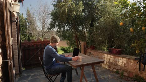 A-man-sitting-at-his-laptop-drinks-coffee-outside-a-rural-house,-with-plants-sorrounding-him