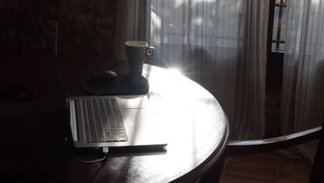 A-shot-of-a-laptop-and-a-steaming-cup-of-coffee-on-a-circular-wooden-table-near-a-window-with-curtains-at-sunrise