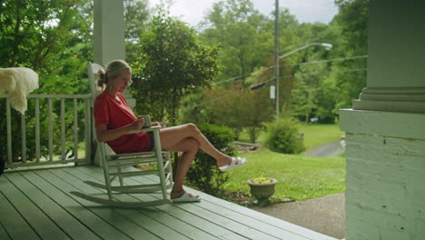 Young-woman-sitting-on-a-rocking-chair-on-her-front-porch,-relaxing-and-drinking-tea