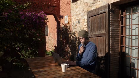 A-man-sitting-at-the-laptop-outside-a-country-house,-touching-his-goatee-whilw-thinking
