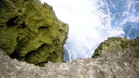 POV-adventurer-looks-over-mossy-green-seacliff-to-see-jagged-rock-and-strong-wave