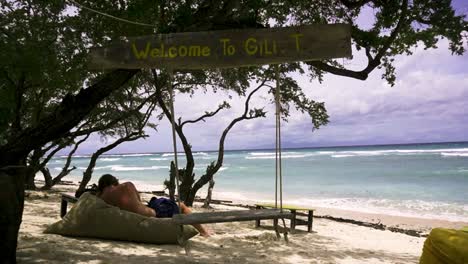 Iconic-welcome-to-Gili-T-sign-with-swing-and-man-lounging-on-bean-bag-oceanside