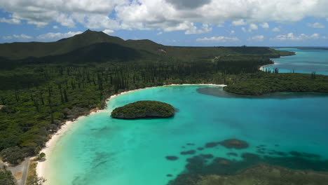 The-white-sand-beach-of-Kanumera-Bay,-Isle-of-Pines-and-a-view-of-the-Sacred-Rock-in-the-lagoon---ascending-aerial-view