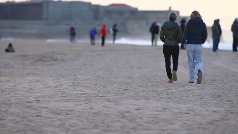 Zwei-Frauen-Gehen-Während-Der-Billabong-Surfmeisterschaft-Am-Strand-Spazieren,-Am-Strand-überfüllt-Mit-Großen-Und-Starken-Wellen-Dahinter,-Strand-Von-Carcavelos,-Cascais,-Portugal