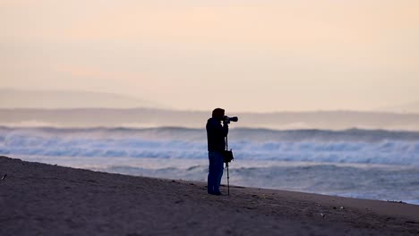 Fotografensilhouette-Am-Strandsonnenaufgang-Mit-Großen,-Verschwommenen-Wellen-Im-Hintergrund