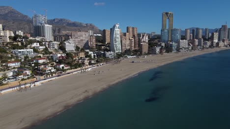 Aerial-descending-shot-showing-people-walking-along-beach-in-Benidorm-during-sunny-day-with-blue-sky-in-summer---Costa-Blanca,Alicante