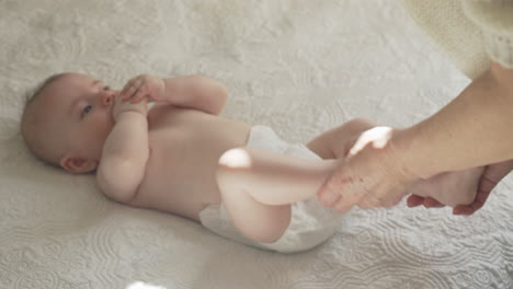 cute-happy-baby-laying-on-his-back,-mother-playing-with-little-legs-in-white-bed-sheets