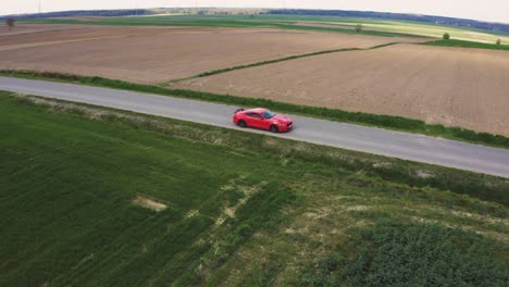 Filmische-Drohnenaufnahme-Eines-Orangefarbenen-Ford-Mustang-GT,-Der-Eine-Gerade-Straße-Entlang-Fährt,-Umgeben-Von-Grünen-Feldern