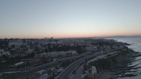 Eine-Atemberaubende-Luftaufnahme-Von-São-Pedro-Do-Estoril-Bei-Sonnenaufgang,-Die-Die-Fahrenden-Züge-Und-Die-Wunderschöne-Küstenstadt-Cascais-In-Portugal-Zeigt