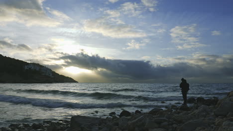 morning-time-lapse-near-rocky-seaside,-mans-silhouette-walks-across,-clouds-moving-fast-sun-rays-breaking