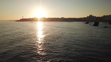 Drohne-Fliegt-über-Schimmerndes-Wasser-Mit-Der-Küste-Von-Biarritz-Und-Dem-Leuchtturm-Im-Hintergrund-Bei-Sonnenuntergang,-Frankreich