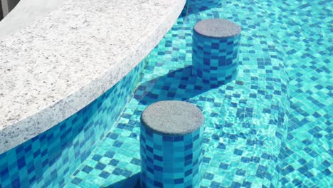 Slow-motion-shot-of-concrete-seats-at-the-side-of-a-pool-bar-with-water-reflecting