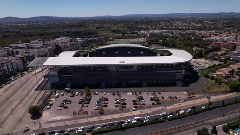Advancing-Aerial-View-of-an-Immense-Stadium-in-the-South-of-France-with-Busy-Road-in-Front