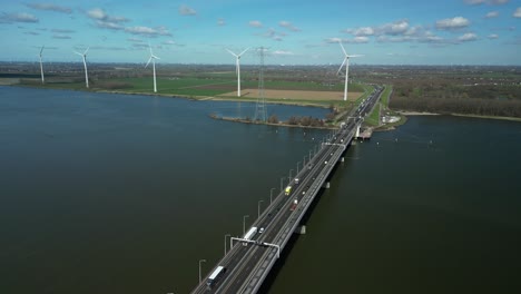 Drone-flies-high-above-a-river-and-a-bridge-for-traffic,-wind-turbines-on-the-horizon