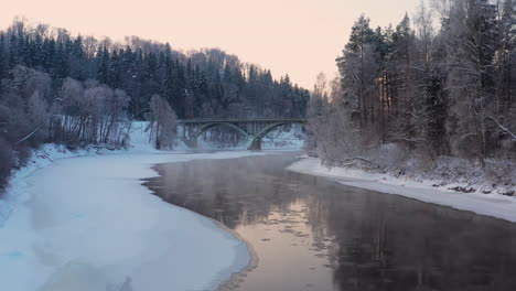 Snow-covered-forest-and-icy-river-with-bridge-at-sunrise,-drone-shot