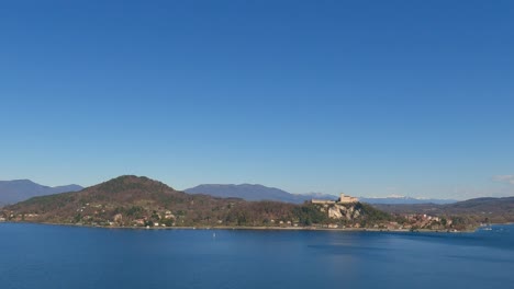 Panoramablick-Auf-Den-Wunderschönen-Lago-Maggiore-Und-Die-Festung-Von-Angera-Mit-Klarem-Himmel-Und-Boot,-Das-über-Ruhiges-Wasser-Segelt