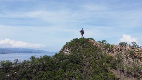 Eine-Aufsteigende-Luftdrohnenaufnahme-Der-Berühmten-Cristo-Rei-Statue-Mit-Der-Stadt,-Dem-Meer-Und-Grünen-Hügeln-Im-Hintergrund-|-Dili,-Timor-Leste