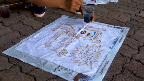 Artist-paints-totem-on-fabric-on-street-sidewalk,-Indonesian-folk-art-close-up