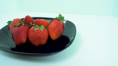 Fresh-big-red-tasty-ripe-strawberries-rotates-slowly-on-a-black-plate-on-light-blue-background,-healthy-food-concept,-medium-shot,-camera-rotate-right