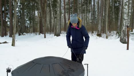 A-young-caucasian-girl-playing-large-steel-drum-on-winter-forest-in-snow-covered-winter-pine-forest-at-sunny-day,-alone-in-frosty-weather,-push-in-gimbal-shot