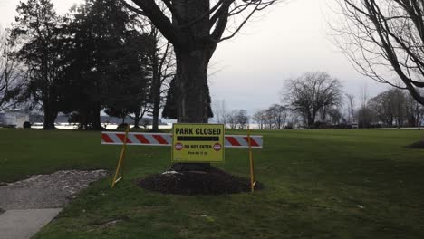 Schilder-„Park-Geschlossen,-Betreten-Verboten“-Schwenken-Seitlich-über-Den-Vordereingang-Des-Parks-Mit-Bäumen-Und-Rasenfläche