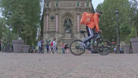 A-slow-motion-shot-of-the-Fontaine-Saint-Michel-in-the-streets-of-Paris,-with-orange-bicylce-rider-and-people-walking-around