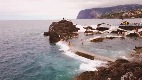 People-Swim-At-Oceanside-Natural-Volcanic-Swimming-Pool-with-Doca-do-Cavacas-Seafood-Restaurant-In-Madeira-Island,-Portugal