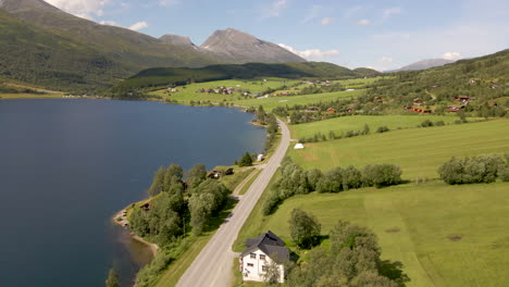 Cars-Driving-On-The-Road-By-The-Lake-Shore-Of-Eidsvatnet-In-Eidsdal,-Norway-With-Mountain-Village-Of-Aarset-In-Background
