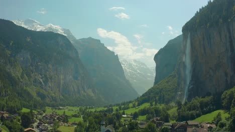 Flug-über-Die-Stadt-Im-Malerischen-Grünen-Tal-Mit-Epischem-Wasserfall,-Schweiz