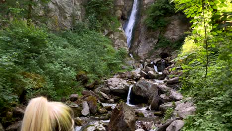 Tilt-up-of-young-woman-take-picture-of-giant-rural-waterfall-in-Austria-during-summer---4K-slow-motion-rear-view