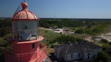 Beautiful-revealing-aerial-view-of-white-painted-steel-lighthouse-with-red-top-located-in-Pape,-Latvia-at-Baltic-sea-coastline-in-sunny-summer-day,-wide-angle-drone-shot-moving-bacwards-close