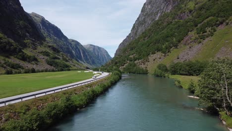 European-road-E39-passing-through-deep-valley-between-glacial-river-and-green-farmland-grass-fields---Aerial-slowly-moving-forward-above-river