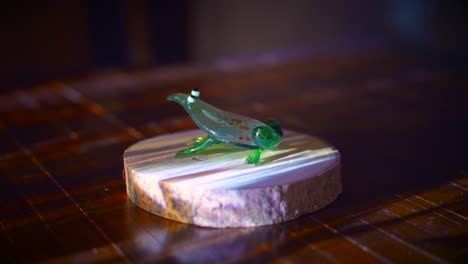 Kunstvolle-Froschfigur-Aus-Mundgeblasenem-Glas,-Inszeniert-Auf-Holzsockel-Mit-Projektorlicht
