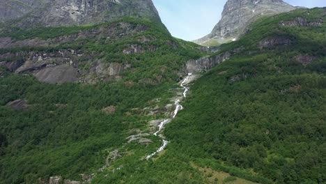 River-flowing-down-hillside-in-Olden-Norway---Tall-mountains-and-lush-forest-with-melted-glacier-water---Norway-aerial