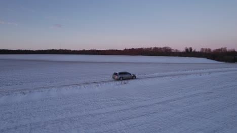 Car-Drives-On-An-Icy-Snow-Covered-Road-In-Denmark,-Scandinavia-Drone-Flying-Beside-A-Car-Driving-On-An-Icy-Road-With-Snow-Covering-The-Fields-With-A-Deep-Blanket-Of-Snow