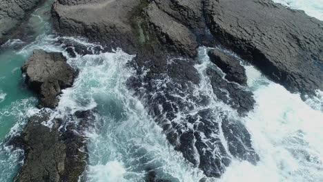 Luftaufnahme,-Die-über-Küstenfelsen-Fliegt-Und-Auf-Die-Wellen-Des-Ozeans-Blickt,-Die-An-Schwarzen-Felsen-Brechen
