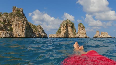Fpv-of-man-legs-and-feet-relaxing-while-floating-on-sea-water-with-Scopello-Stacks-or-Faraglioni-in-background