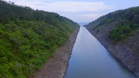 Aerial-backwards-shot-of-small-river-dam-surrounded-by-rocky-shore-and-green-plants-growing-on-hill