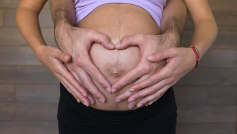 Expecting-parents-form-a-heart-with-hands-on-pregnant-woman-belly,-close-up