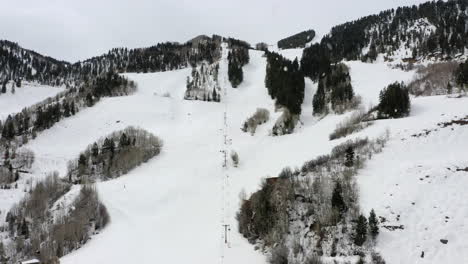 Cable-cart-lift-rising-up-tall-skiing-hill-slope-during-snowfall,-aerial-drone-view