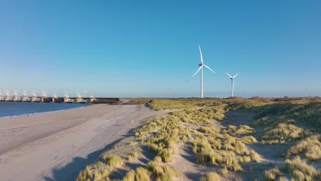 Aerial-shot-flying-over-dunes-and-a-beach-towards-wind-turbines-and-the-Eastern-Scheldt-storm-surge-barrier-in-Zeeland,-the-Netherlands,-on-a-beautiful-sunny-day
