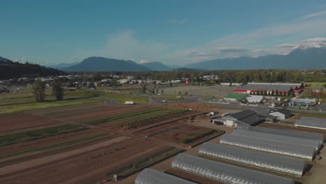Local-Harvest-farm-in-Chilliwack-BC-brown-fields-freshly-planted-crops-in-spring-Wide-aerial-reversing-revealing-traffic-trees-mountains-valley-clouds-blue-sky-sunny-day