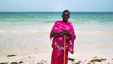 Native-african-man-in-pink-clothing-holding-stick-stands-on-sand-beach