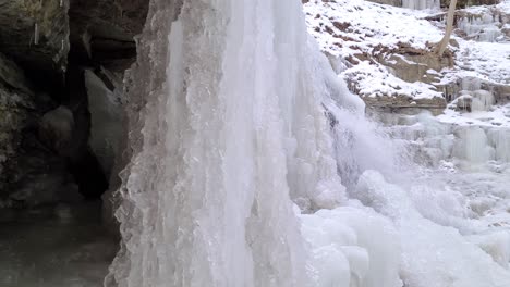 Halve-Frozen-Waterfall-Flowing-From-a-Mountain-at-Winter-Time,-Icy-Ground---Slow-Sliding-Reveal-Shot