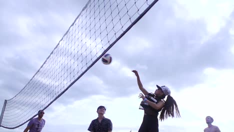 Asiatische-Frau-Mit-Mütze-Schlägt-Volleyball-über-Netz-Während-Unisex-Team-Volleyballwettbewerb-Am-Strand,-Gefilmt-In-Zeitlupe