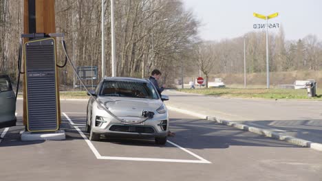Handsome-businessman-using-smartphone-and-leans-on-luxurious-vehicle-while-waiting-for-electric-car-to-finish-charging-next-to-the-highway---Leuven,-Belgium