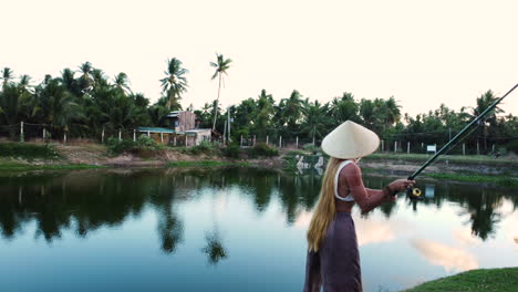 Attractive-woman-with-conical-hat-fishing-in-tropical-Vietnam-lake,-orbit-view