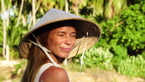 Cute-woman-with-conical-hat-in-tropical-forest-smiles-against-sunshine,-close-up-view
